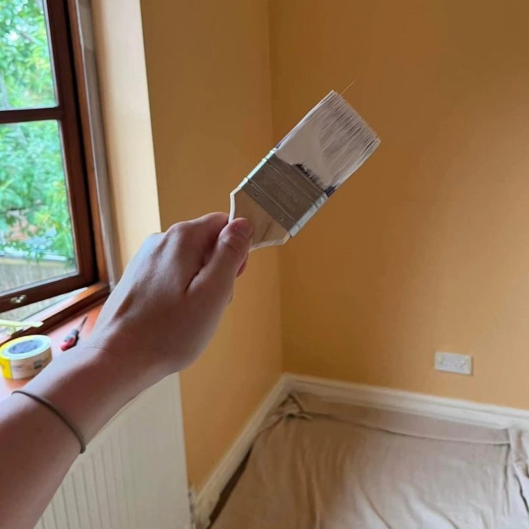 A hand holds a paintbrush in a room with yellow walls and a covered floor.