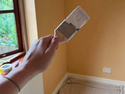 A hand holding a paintbrush in a room with yellow walls and a window.