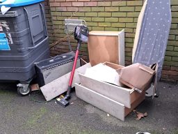 Discarded furniture and items, including a vacuum cleaner, near a waste bin.