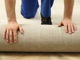 Person kneeling, unrolling a beige carpet on a wooden floor.